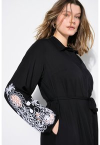 Schwarzes Kleid mit Kragen zum Knöpfen, langen Ärmeln mit weißen gestickten Mustern und einem gebundenen Taillenbereich für eine figurbetonte Silhouette.
