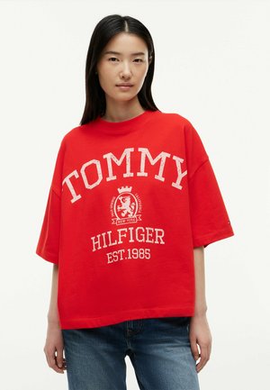 Junge Frau trägt ein locker sitzendes rotes Tommy Hilfiger T-Shirt mit weißem Logo und blauen Jeans vor einem einfarbigen weißen Hintergrund.