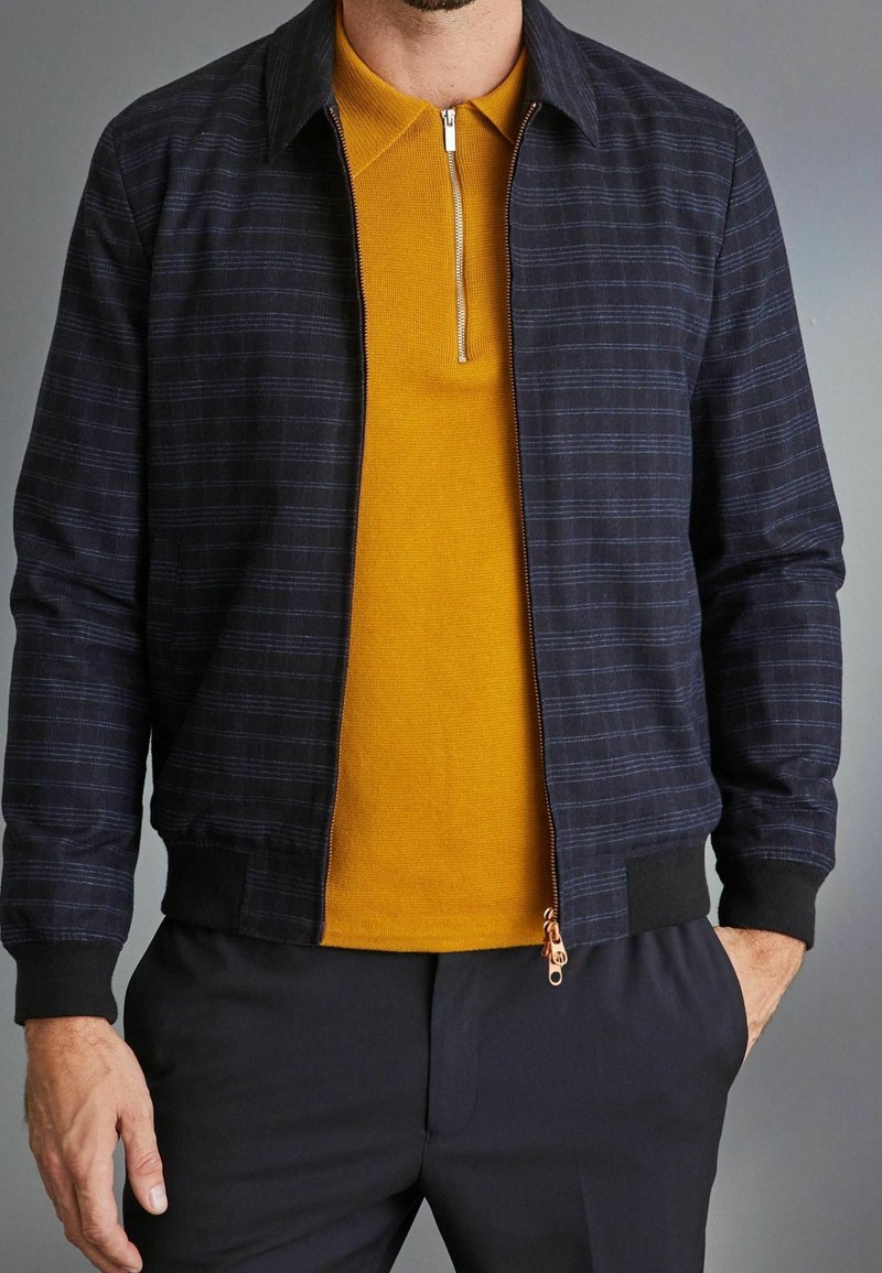 Navy checkered bomber jacket with ribbed cuffs, paired with a yellow zip-neck polo shirt, featuring a textured fabric and slim fit.