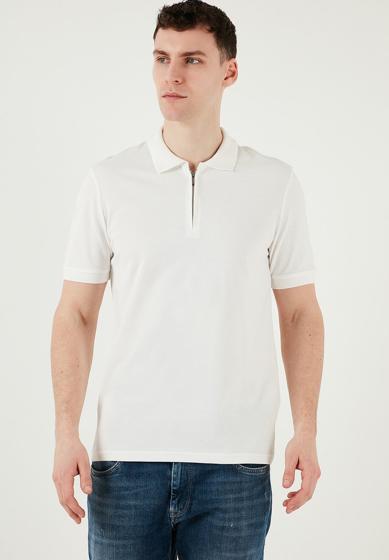 Vit polotröja med en fjärdedels dragkedja, korta ärmar och texturerad krage; matchad med blå jeans. Slät tyg och figursydd design.