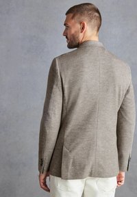 Braun gemusterter Blazer mit taillierter Passform, ausgestattet mit einem Schlitz und zwei Seitentaschen, aus strukturiertem Stoff gefertigt.