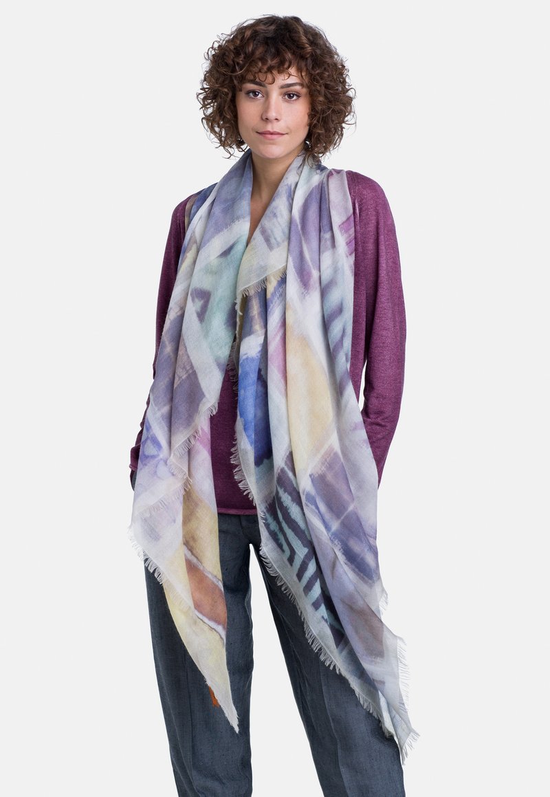 Scarf leggero e multicolore con un motivo geometrico e bordi sfrangiati, indossato sopra una maglietta viola e pantaloni grigi. Texture morbida, design fluido.