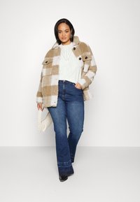 Casaco de fleece xadrez em bege e branco com grandes botões pretos, combinado com uma blusa clara e jeans azul largas. A segurar uma bolsa trançada branca.