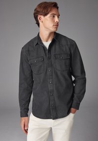 Camicia di denim nera con spalle a punta, due tasche sul petto e chiusura a bottoni. Indossata sopra una maglietta grigia chiara, abbinata a pantaloni bianchi.