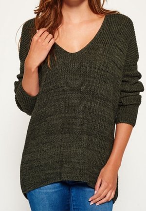 Ciemnozielony dzianinowy sweter z dekoltem w serek i długimi rękawami, noszony z niebieskimi dżinsami, widoczny od szyi do bioder.