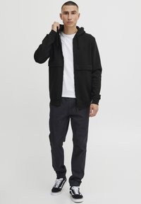 Sudadera negra con cremallera y bolsillos frontales, diseño en capas, camiseta blanca debajo, combinada con pantalones oscuros ajustados y zapatillas negras.