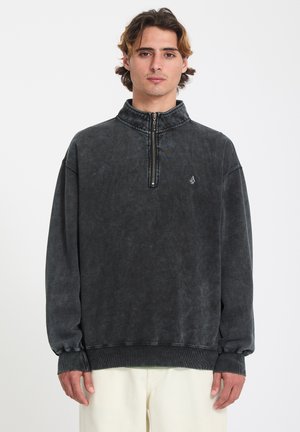 Jeune homme aux cheveux châtain clair portant un sweat-shirt noir délavé à fermeture éclair quart et un pantalon beige clair, debout devant un fond blanc uni.