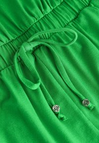 Tissu en coton vert avec une taille élastique et un cordon de serrage. Présente des accents métalliques et une texture lisse. Design léger et décontracté.