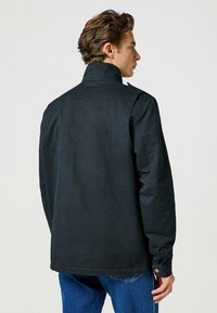 Schwarze Canvasjacke mit hohem Kragen, geradem Schnitt, Knopfverschluss und sichtbaren Nähten an Schultern und Rücken.