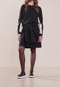 Czarna bluza z białą i czerwoną grafiką gwiazdy na rękawach, nałożona na czarną spódnicę z detalem w postaci wiązania, w zestawieniu z błyszczącymi czarnymi butami.