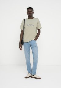 Ανοιχτό λαδί βαμβακερό μπλουζάκι με μπλε λογότυπο, σε συνδυασμό με ανοιχτό μπλε τζιν παντελόνι και μπεζ αθλητικά παπούτσια. Το μοντέλο στέκεται μπροστά σε λευκό φόντο.