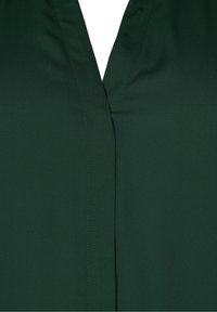 Blusa verde con scollo a V, texture liscia, leggero luccichio e dettagli cuciti lungo il pattina. Nessun motivo o hardware visibile.