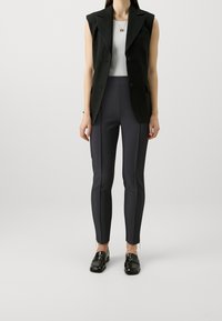 Blazer sans manches noir sur un haut côtelé blanc, associé à un pantalon ajusté gris foncé avec des zips sur le côté et des mocassins noirs brillants.