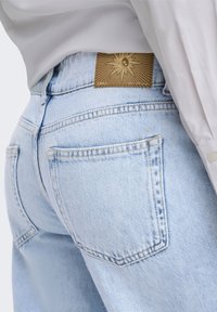 Jean en denim bleu clair avec des poches arrière droites et une étiquette en cuir marron affichant un motif soleil et lune, porté avec une chemise blanche.