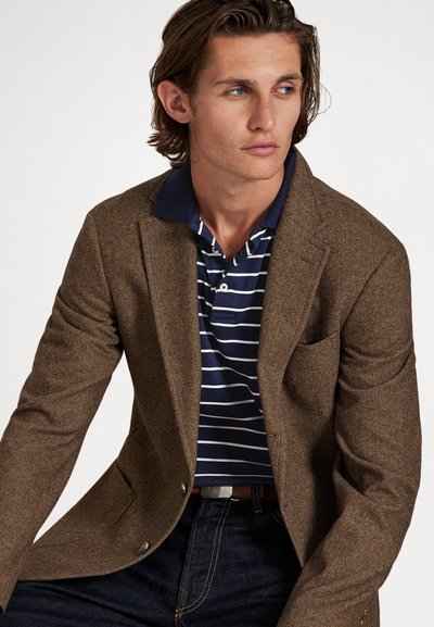 Jauns vīrietis ar brūnu zivju kaulu raksta žaketi pār plecu melni zilas un baltas svītrains polo krekls, sēžot un raugoties uz sāniem pret balta fona.