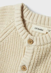 Beige stickad tröja med ribbad krage, prydd med träknappar och en texturerad mönstring. Etiketten läser "Lil'Atelier."