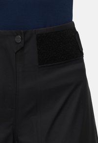 Zwarte broek met een gladde textuur, zij-velcroaccenten en een zilveren knoop op de tailleband, met daarboven een contrasterende blauwe stof.