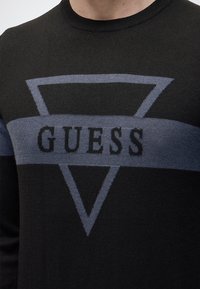 Pull noir avec un grand triangle gris et une bande horizontale portant le mot « GUESS » sur la poitrine.
