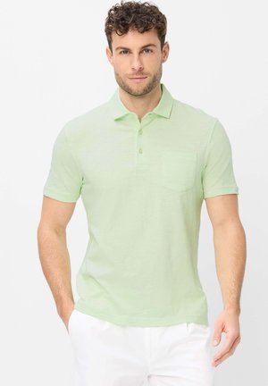 Mann mit lockigem Haar, der ein hellgrünes kurzärmeliges Polohemd und weiße Hose trägt, steht vor einem schlichten weißen Hintergrund.