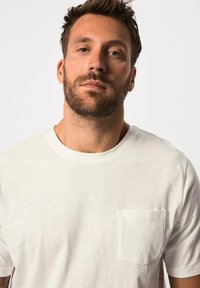 Camiseta de algodón blanca con cuello redondo y mangas cortas. Presenta un pequeño bolsillo en el pecho y una tela suave, ligeramente texturizada.