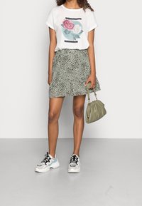 Camiseta blanca con estampado floral, combinada con una falda verde con un patrón de manchas negras. Los accesorios incluyen un bolso verde claro y zapatillas.