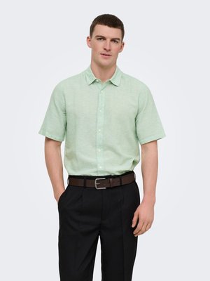 Jeune homme portant une chemise à manches courtes vert clair boutonnée, rentrée dans un pantalon noir avec une ceinture marron, debout avec une main dans la poche.