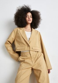 Blazer beige coupé avec des revers crantés et des poches avant, accompagné d'un pantalon taille haute assorti avec des coutures blanches contrastées.