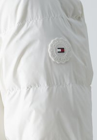 Doudoune blanche à texture matelassée, arborant un patch circulaire gris avec "Tommy Hilfiger" et un petit emblème en rouge, blanc, et bleu.