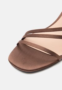 Καφέ σατέν σανδάλι με λουράκια, τετράγωνη μύτη και τρεις λεπτές λωρίδες στο μπροστινό μέρος σε ανοιχτό φόντο.