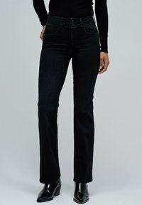 Mujer con vaqueros negros de tiro alto y patas rectas, y botas de tacón negras hasta el tobillo, de pie contra un fondo gris claro uniforme.