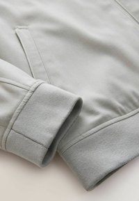 Gaiši pelēks sporta krekls ar gludu tekstūru, ribinātām piedurknēm un sānu kabatu. Dizains ir vienkāršs ar tīrām līnijām un minimāliem akcentiem.