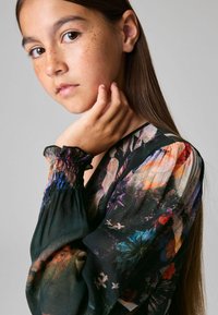 Blus i genomskinligt svart tyg med blommönster, långa puffärmar och elastiska volangmuddar. Modellen posar med handen nära ansiktet och visar detaljerna.