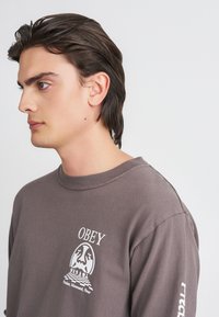 Siva dugouga majica dugih rukava od mekog materijala s bijelim grafičkim prikazom i natpisom "OBEY" te jedinstvenim dizajnom na prsima i rukavima.
