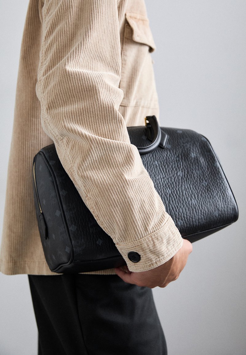 Borsa in pelle nera con superficie texture, tenuta da una persona in una camicia di corduroy chiara. Presenta una forma curva e una maniglia superiore.