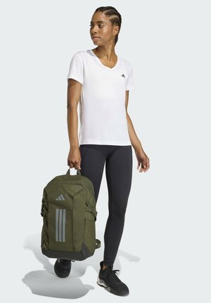 Mujer con camiseta blanca y leggings negros sosteniendo una mochila verde oliva con el logo reflectante de adidas, mirando hacia un lado.