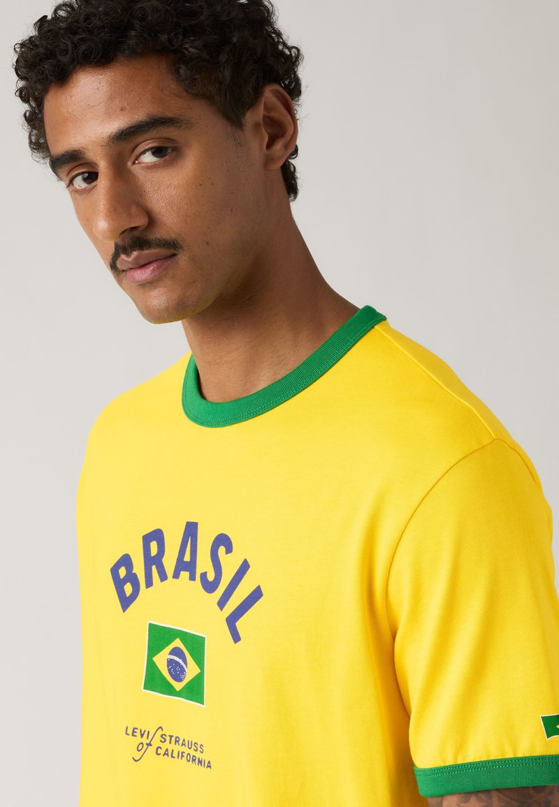 Homme avec une moustache portant un t-shirt jaune avec un liseré vert, le texte "BRASIL" et un drapeau brésilien sur la poitrine et la manche.