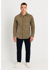 Olivgrüne, wildlederähnliche Hemdjacke mit Knopfverschluss, zwei Brusttaschen und Kragen, kombiniert mit marineblauen Hosen und weißen Sneakers.