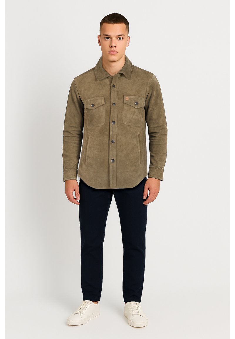 Olivgrüne, wildlederähnliche Hemdjacke mit Knopfverschluss, zwei Brusttaschen und Kragen, kombiniert mit marineblauen Hosen und weißen Sneakers.