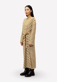 Gestreepte bruine en crèmekleurige langemouwen jurk met een striktaille en een rechte snit. Model draagt zwarte enkellaarzen. Glad, lichtgewicht stof.