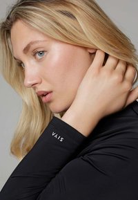 Zwarte langslavige kleding met een strakke pasvorm, soepele textuur en een wit logo "VAIS" op de manchet. Close-up van het gezicht en de hand van een persoon.