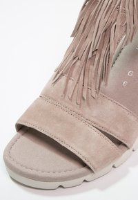Sandale en daim beige avec deux larges brides, détail à franges et semelle rembourrée avec dessous blanc texturé.