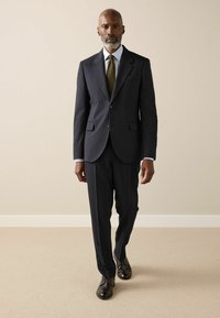 Marineblauer Anzug mit maßgeschneiderter Passform, einreihiger Jacke, Revers mit Kerbe, kombiniert mit einem hellblauen Hemd und einer olivgrünen Krawatte. Braune Lederschuhe.