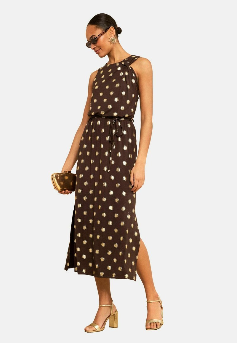 Frau trägt ärmelloses braunes Midikleid mit goldenen Sonnenmustern, goldene Sandalen mit hohen Absätzen, goldene Clutch, Sonnenbrille und sternförmige Ohrringe.