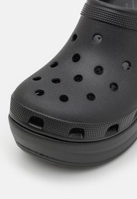 Črne klompe iz teksturirane plastike, ki imajo več ventilacijskih lukenj in rahlo dvignjen, zaobljen prst. Protizdrsna podlaga.