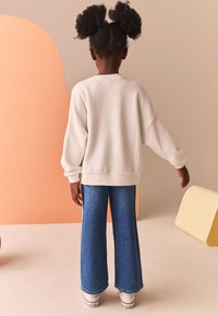 Felpa bianca oversize con spalle scese, abbinata a jeans denim blu a gamba larga. Scarpe casual con suole bianche visibili.