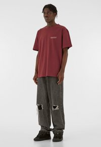 Burgundy oversized t-shirt with short sleeves and a small graphic. Worn with distressed grey jeans and black sneakers. Smooth fabric texture.