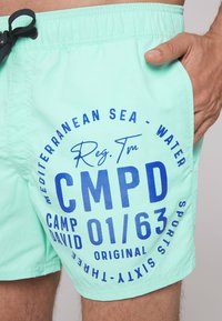 Türkiz színű strandnadrág könnyű anyagból. Kék nyomott szöveg található rajta, beleértve a "CMPD" és "01/63" feliratokat. Rugalmas derékpánt húzózsinórral.