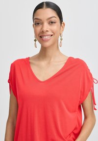 Rode V-hals top met korte mouwen en trekkoordaccenten. Zachte stof, losse pasvorm, met een gladde textuur. Gouden en roze oorbellen.