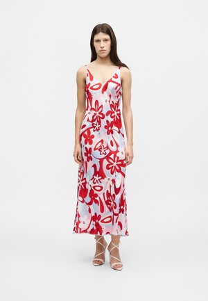 Nő ujjatlan, V-nyakú, bokáig érő ruhában, piros, fehér és világoskék virágos és absztrakt mintával, semleges arckifejezéssel áll.