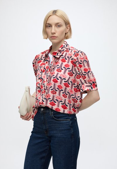 Femme aux cheveux blonds courts portant un chemisier fleuri rouge et rose à motifs et un jean bleu foncé, tenant un petit sac à main beige.